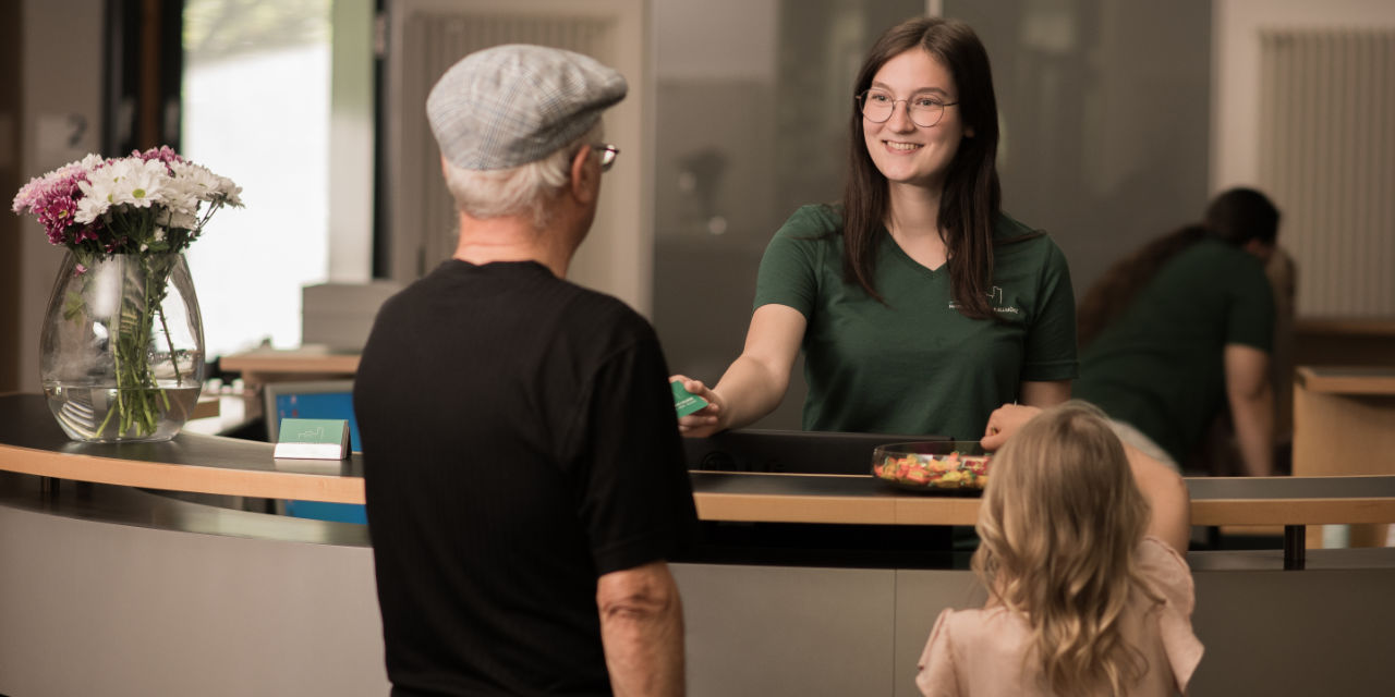 Älterer Patient steht mit Kind an der Rezeption und bekommt dort eine Visitenkarte.